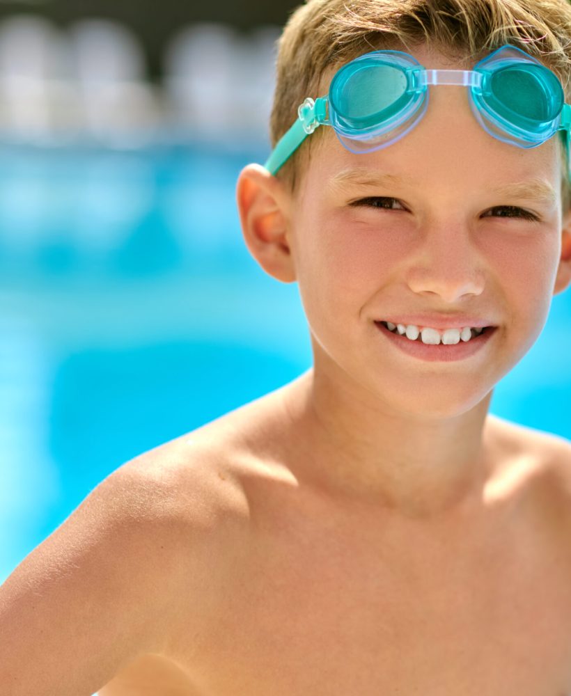 Confidence. School age boy with swimming goggles looking confidently at camera standing near pool on summer day